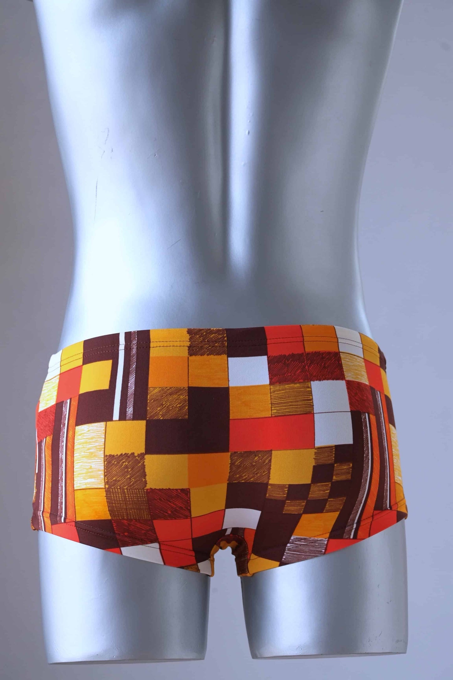 A plastic mannequin wearing a pair of Vintage Tropic swim briefs featuring a retro checkered pattern in orange, red, brown, and white, photographed from the back.
