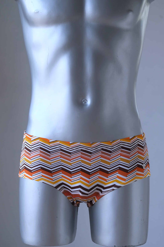 A mannequin wearing a pair of Tropic Vintage swim briefs featuring a brown, orange and yellow chevron pattern, shown from the front.
