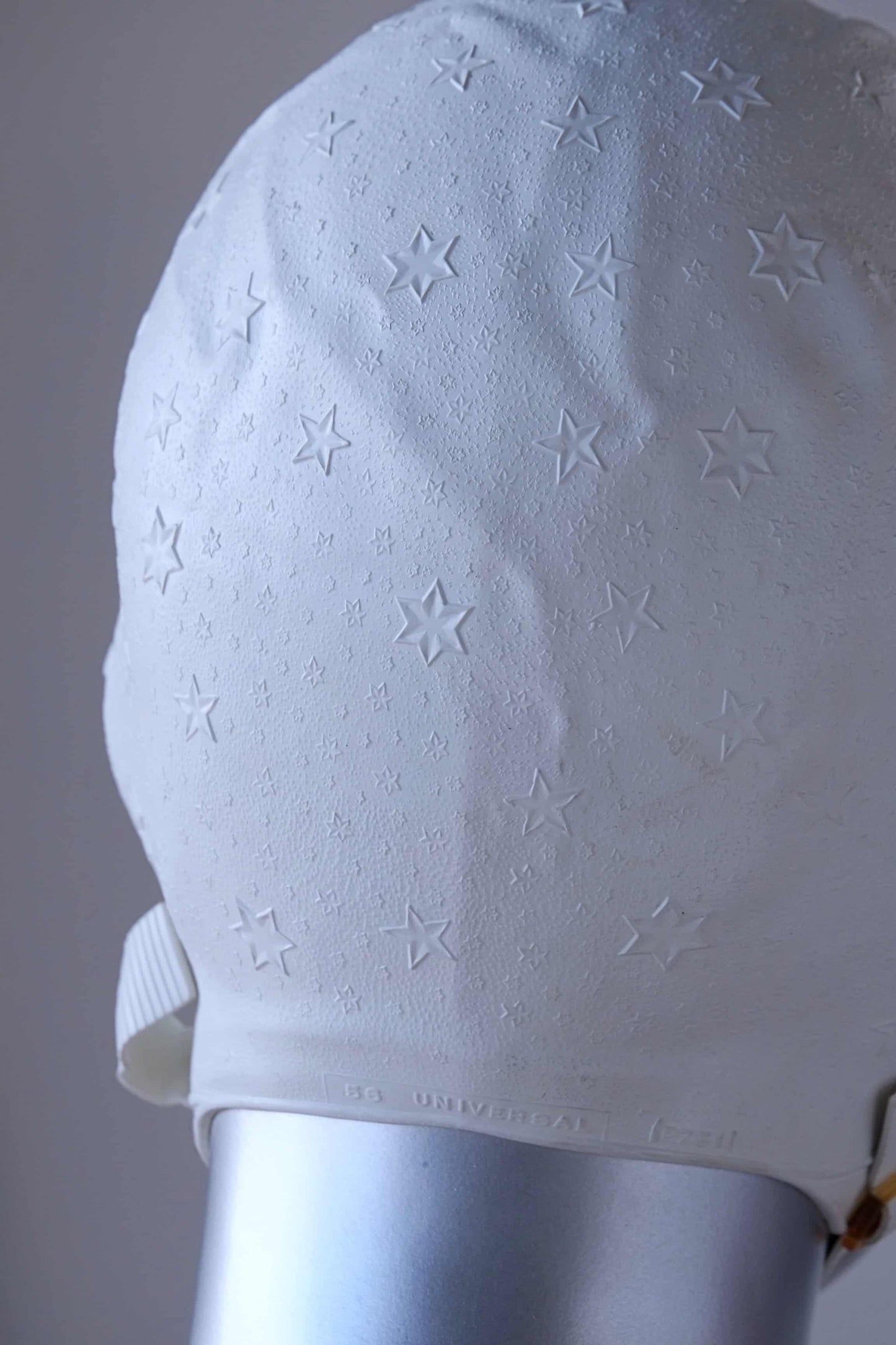 Close up of a vintage 1960s white swim cap made of textured rubber with an embossed stars pattern, worn by a silver mannequin. The cap features a secure chin strap with metal clasps. The mannequin is set against a neutral background.