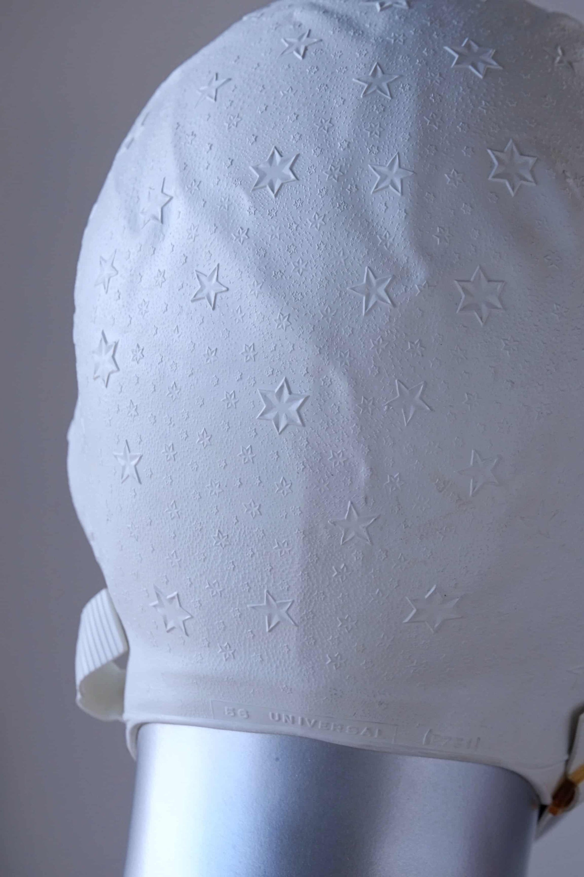 Close up of a vintage 1960s white swim cap made of textured rubber with an embossed stars pattern, worn by a silver mannequin. The cap features a secure chin strap with metal clasps. The mannequin is set against a neutral background.