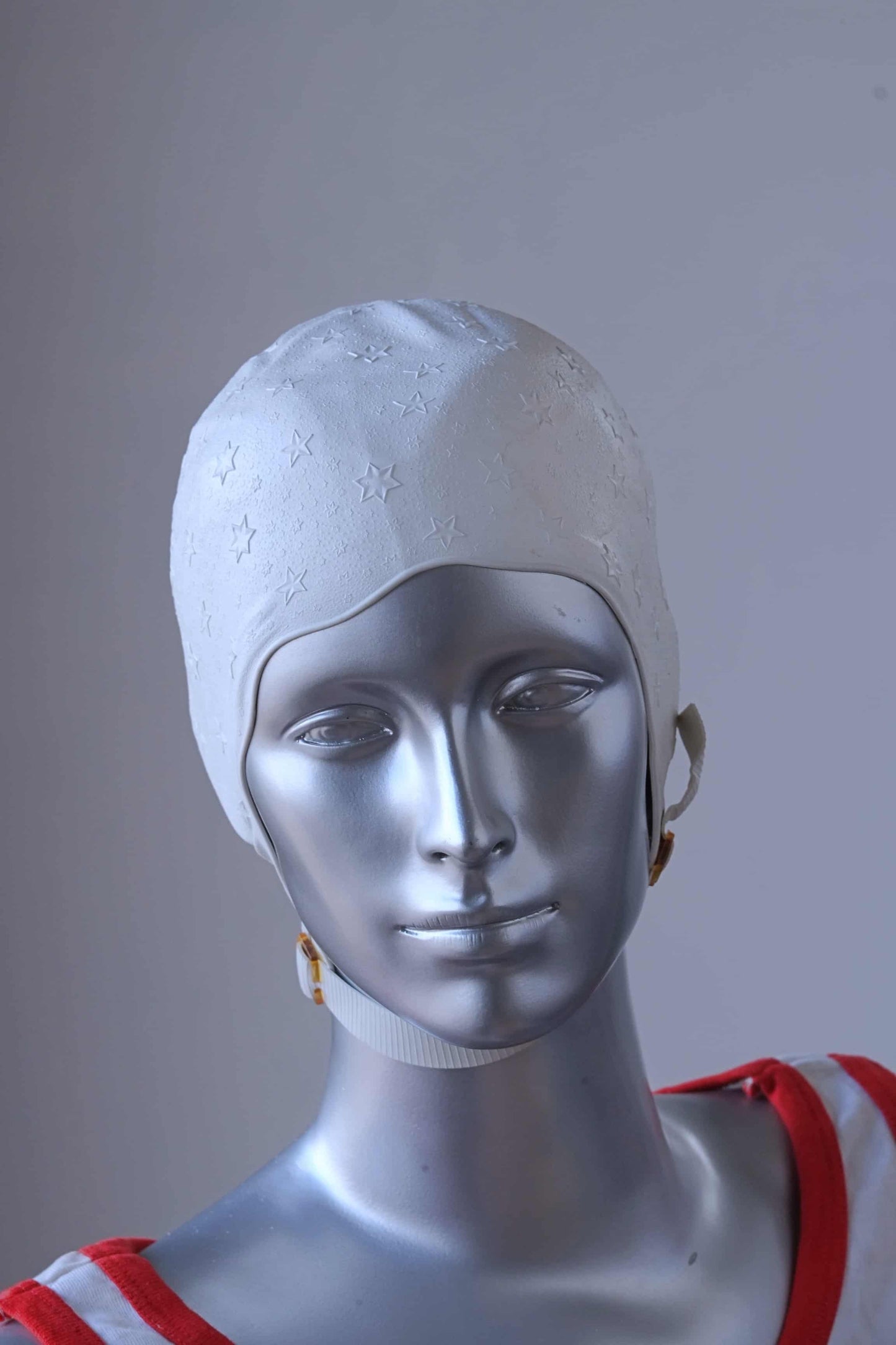 A vintage 1960s white swim cap made of textured rubber with an embossed stars pattern, worn by a silver mannequin. The cap features a secure chin strap with metal clasps. The mannequin is set against a neutral background.