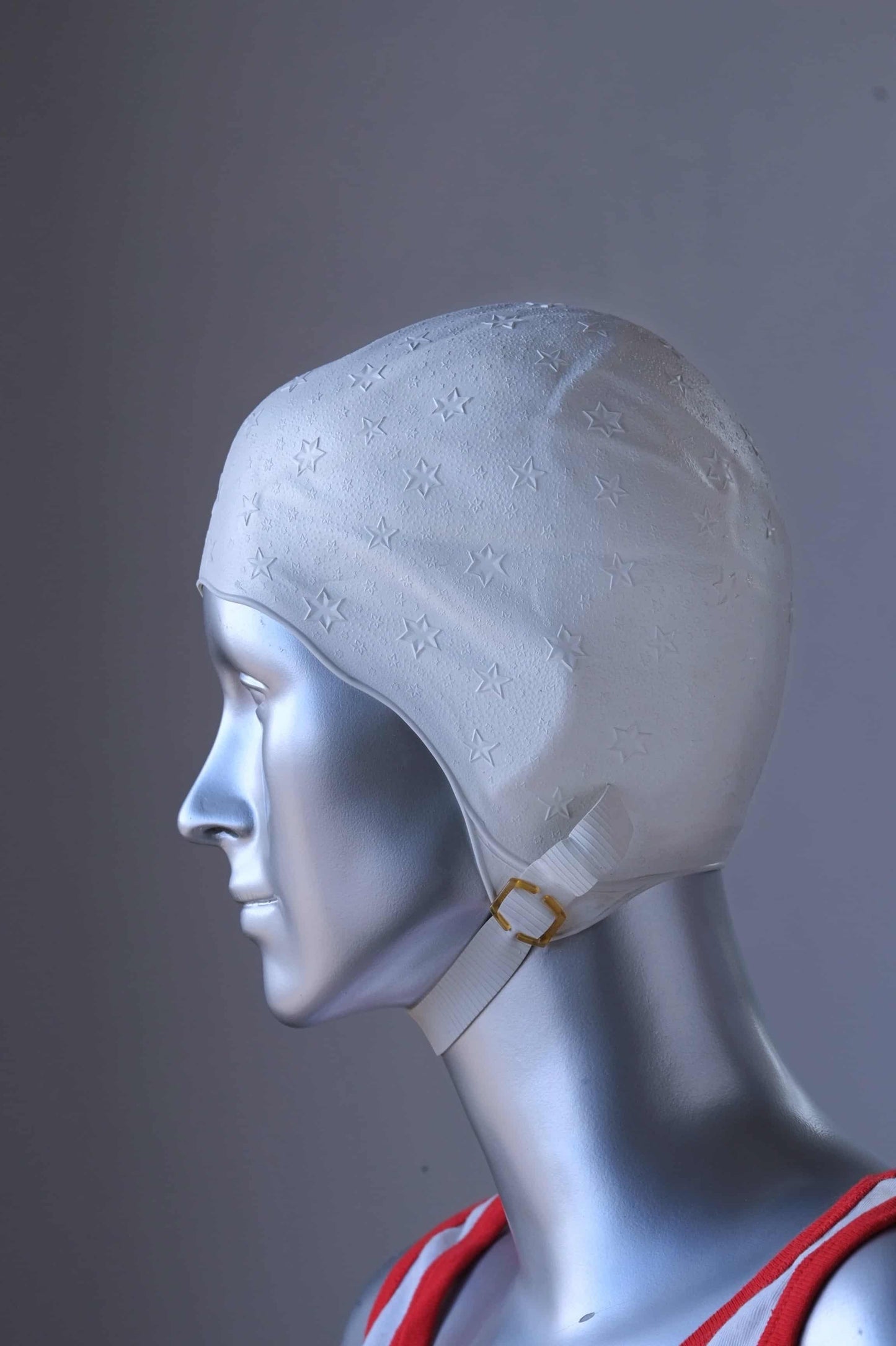Side view of a vintage 1960s white swim cap made of textured rubber with an embossed stars pattern, worn by a silver mannequin. The cap features a secure chin strap with metal clasps. The mannequin is set against a neutral background.