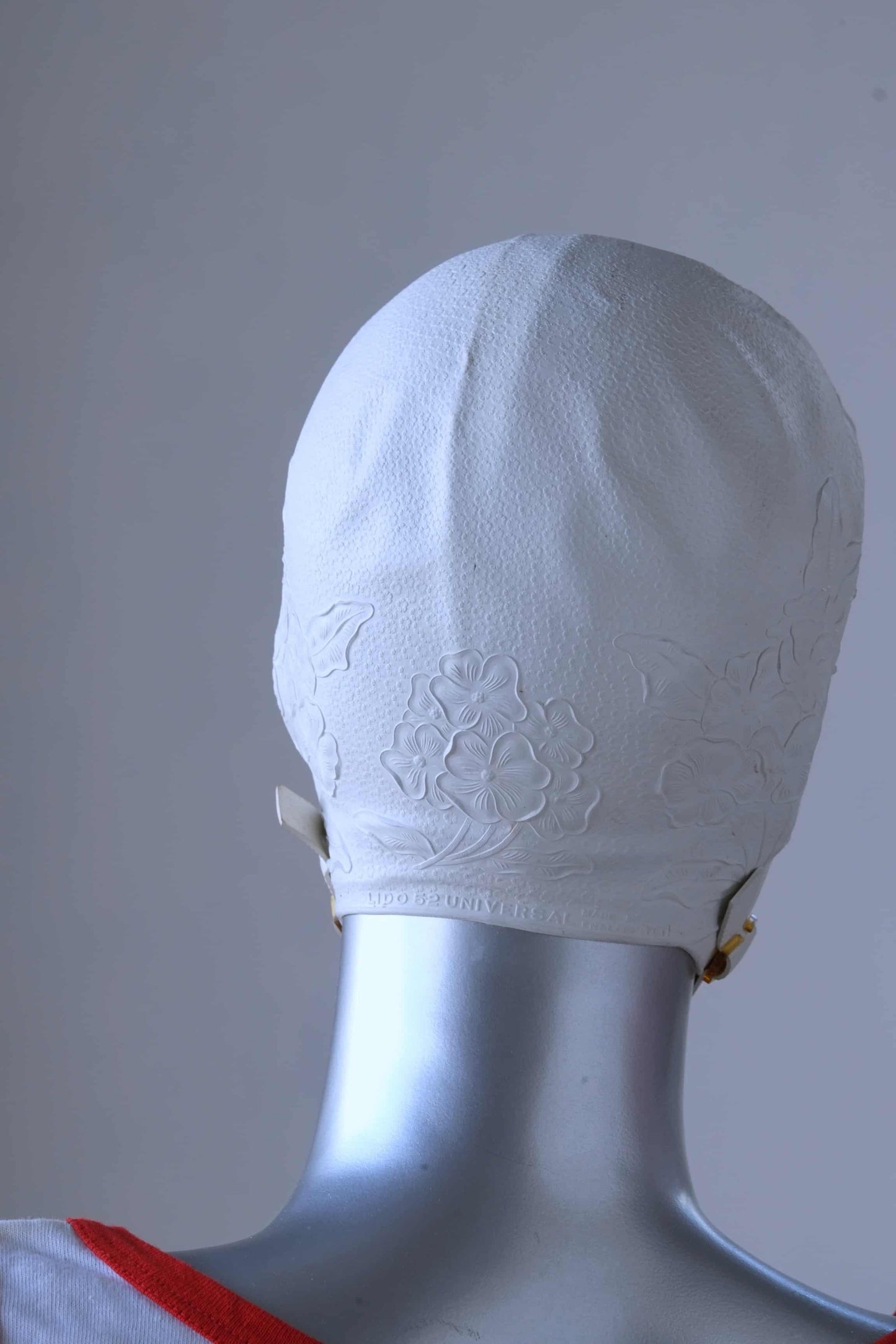Back view of a vintage 1960s white swim cap made of textured rubber with an embossed floral pattern, worn by a silver mannequin. The cap features a secure chin strap with metal clasps. The mannequin is set against a neutral background.