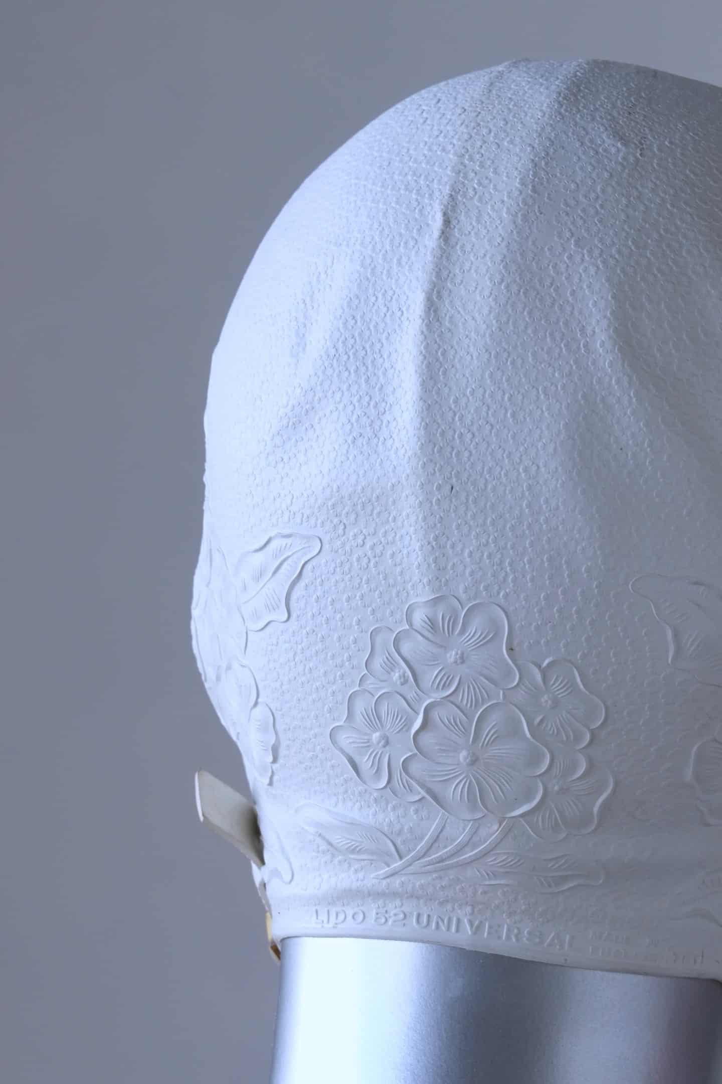 Close up of a vintage 1960s white swim cap made of textured rubber with an embossed floral pattern, worn by a silver mannequin. The cap features a secure chin strap with metal clasps. The mannequin is set against a neutral background.
