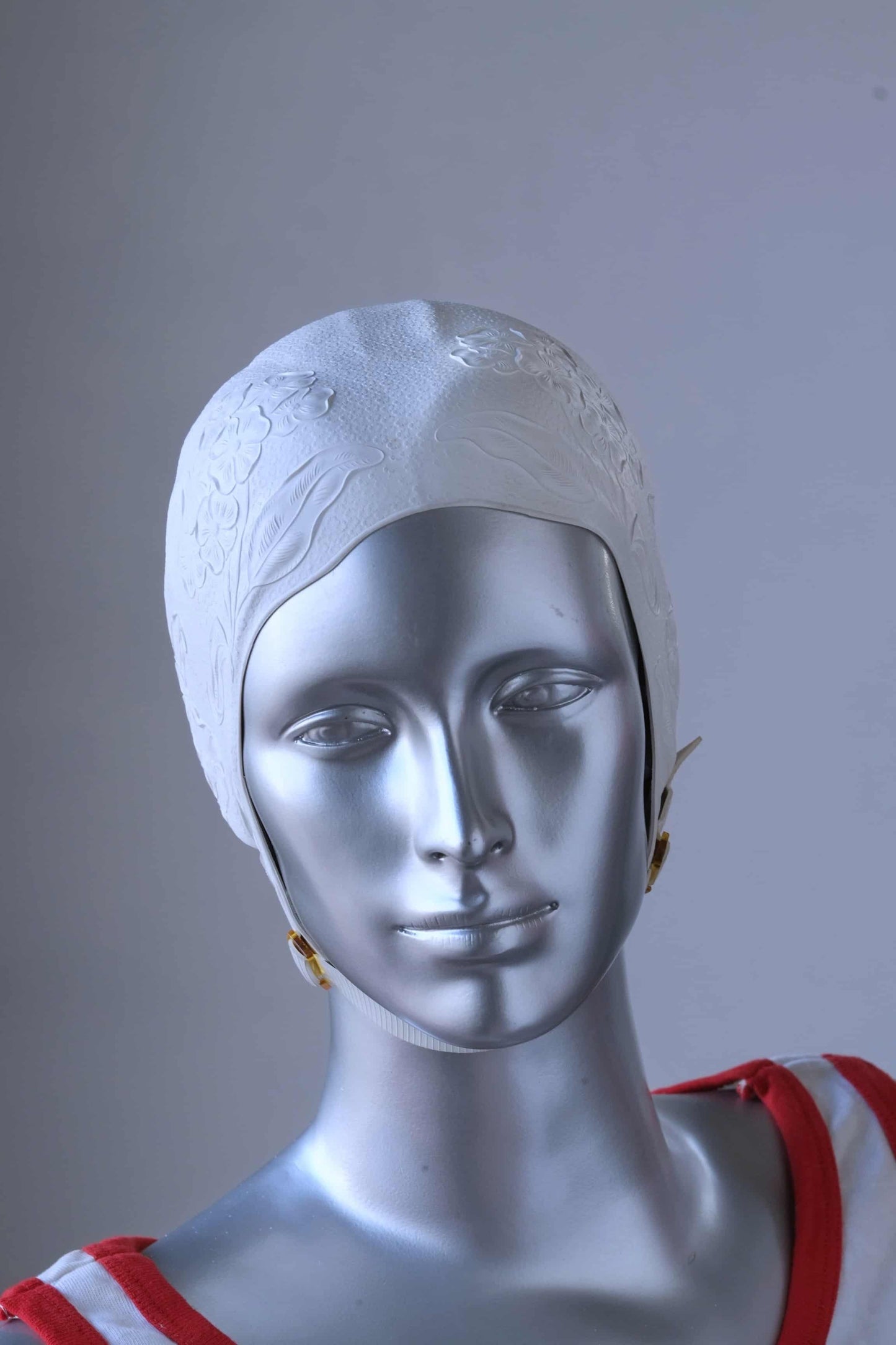 A vintage 1960s white swim cap made of textured rubber with an embossed floral pattern, worn by a silver mannequin. The cap features a secure chin strap with metal clasps. The mannequin is set against a neutral background.