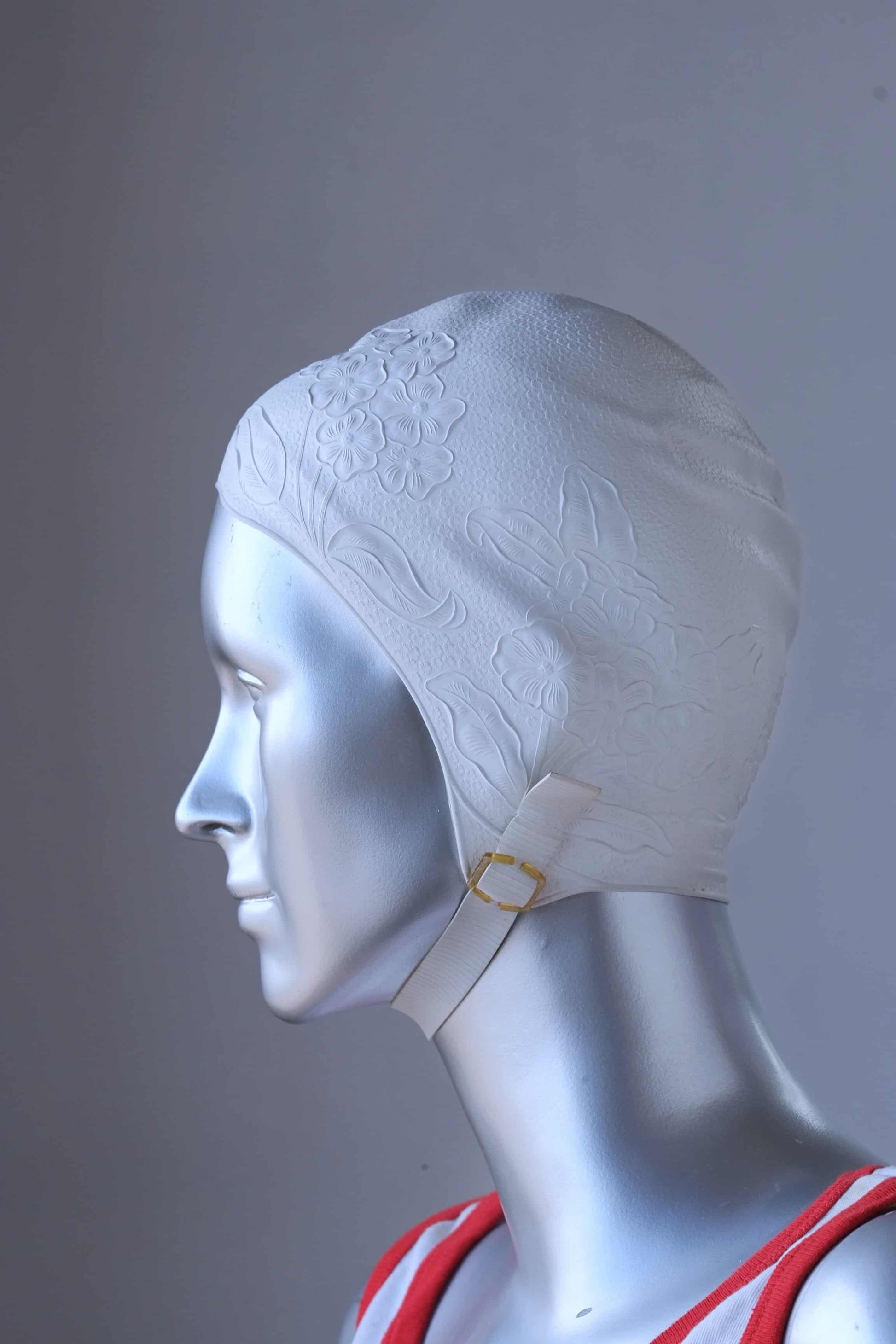 Side view of a vintage 1960s white swim cap made of textured rubber with an embossed floral pattern, worn by a silver mannequin. The cap features a secure chin strap with metal clasps. The mannequin is set against a neutral background.