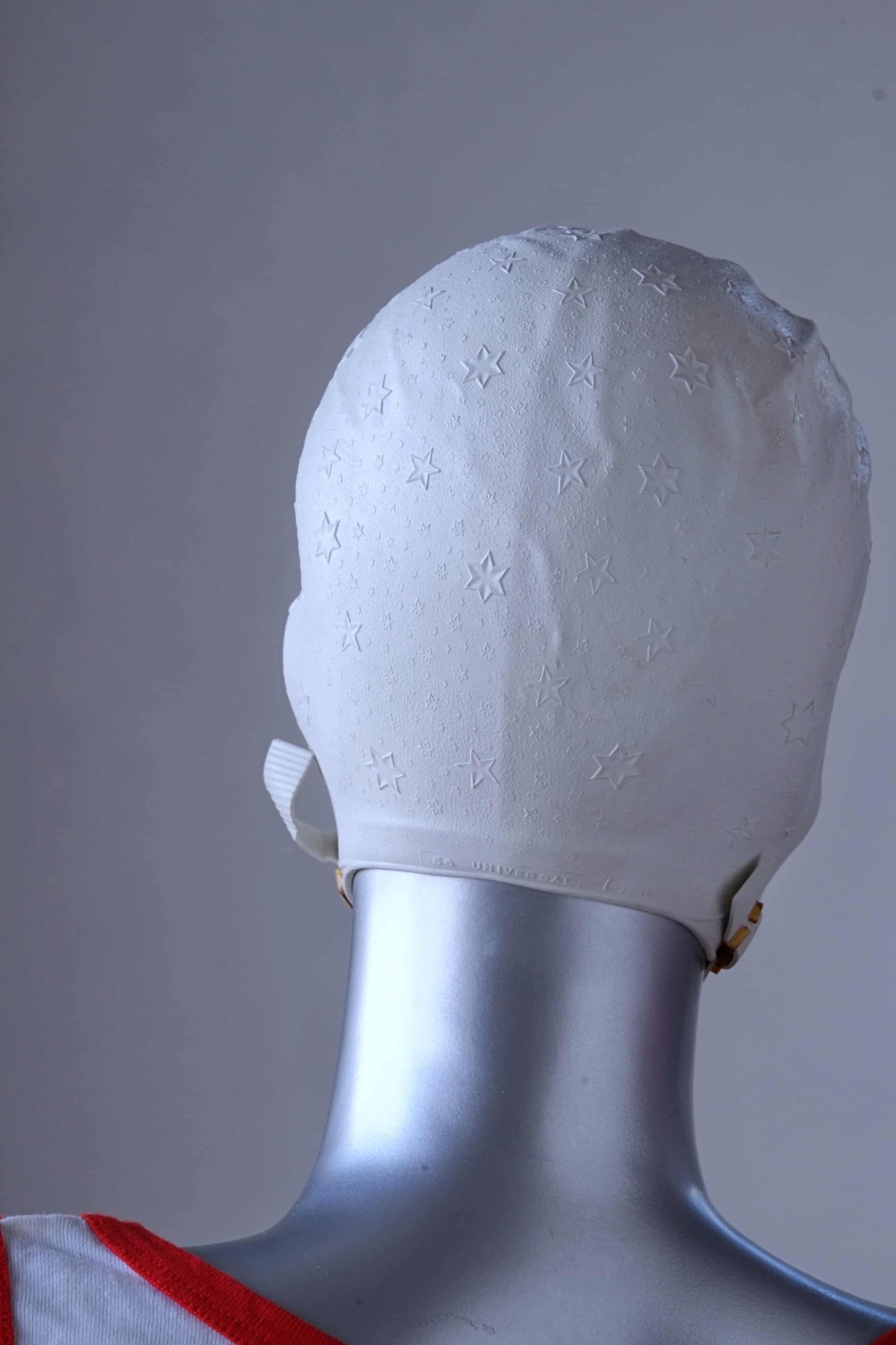 Back view of a vintage 1960s white swim cap made of textured rubber with an embossed stars pattern, worn by a silver mannequin. The cap features a secure chin strap with metal clasps. The mannequin is set against a neutral background.