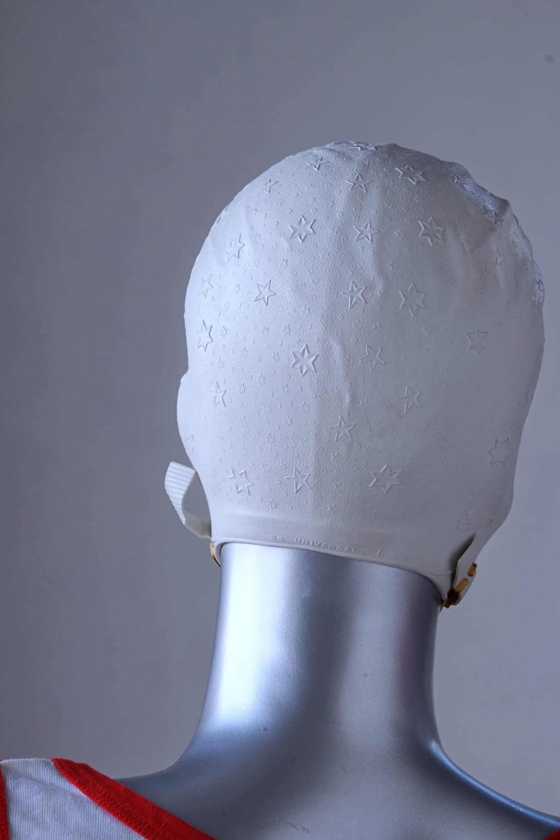 Back view of a vintage 1960s white swim cap made of textured rubber with an embossed stars pattern, worn by a silver mannequin. The cap features a secure chin strap with metal clasps. The mannequin is set against a neutral background.
