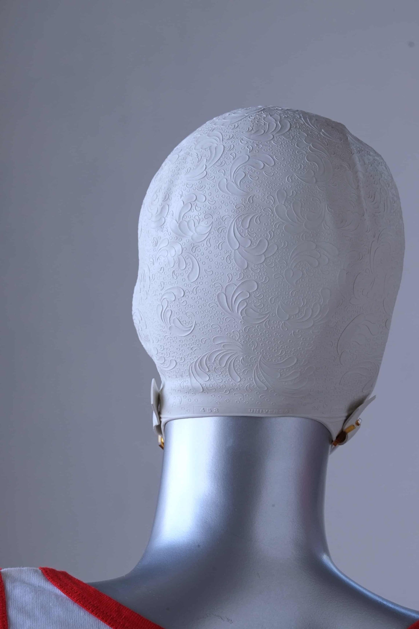 Back view of a vintage 1960s white swim cap made of textured rubber with an embossed waves pattern, worn by a silver mannequin. The cap features a secure chin strap with metal clasps. The mannequin is set against a neutral background.