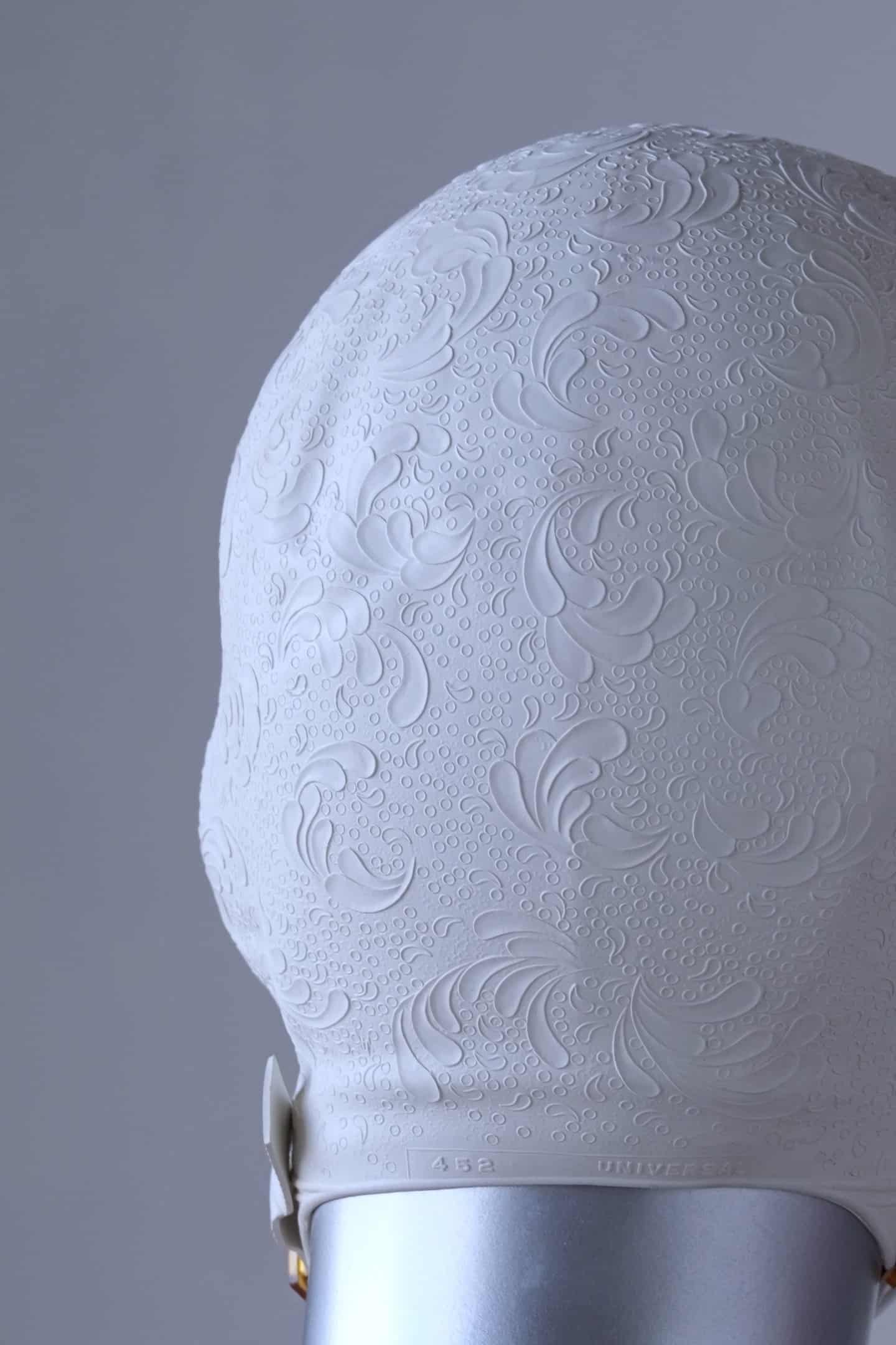Close up of a vintage 1960s white swim cap made of textured rubber with an embossed waves pattern, worn by a silver mannequin. The cap features a secure chin strap with metal clasps. The mannequin is set against a neutral background.