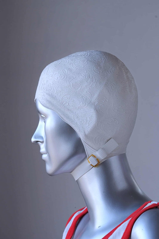 Side view of a vintage 1960s white swim cap made of textured rubber with an embossed waves pattern, worn by a silver mannequin. The cap features a secure chin strap with metal clasps. The mannequin is set against a neutral background.