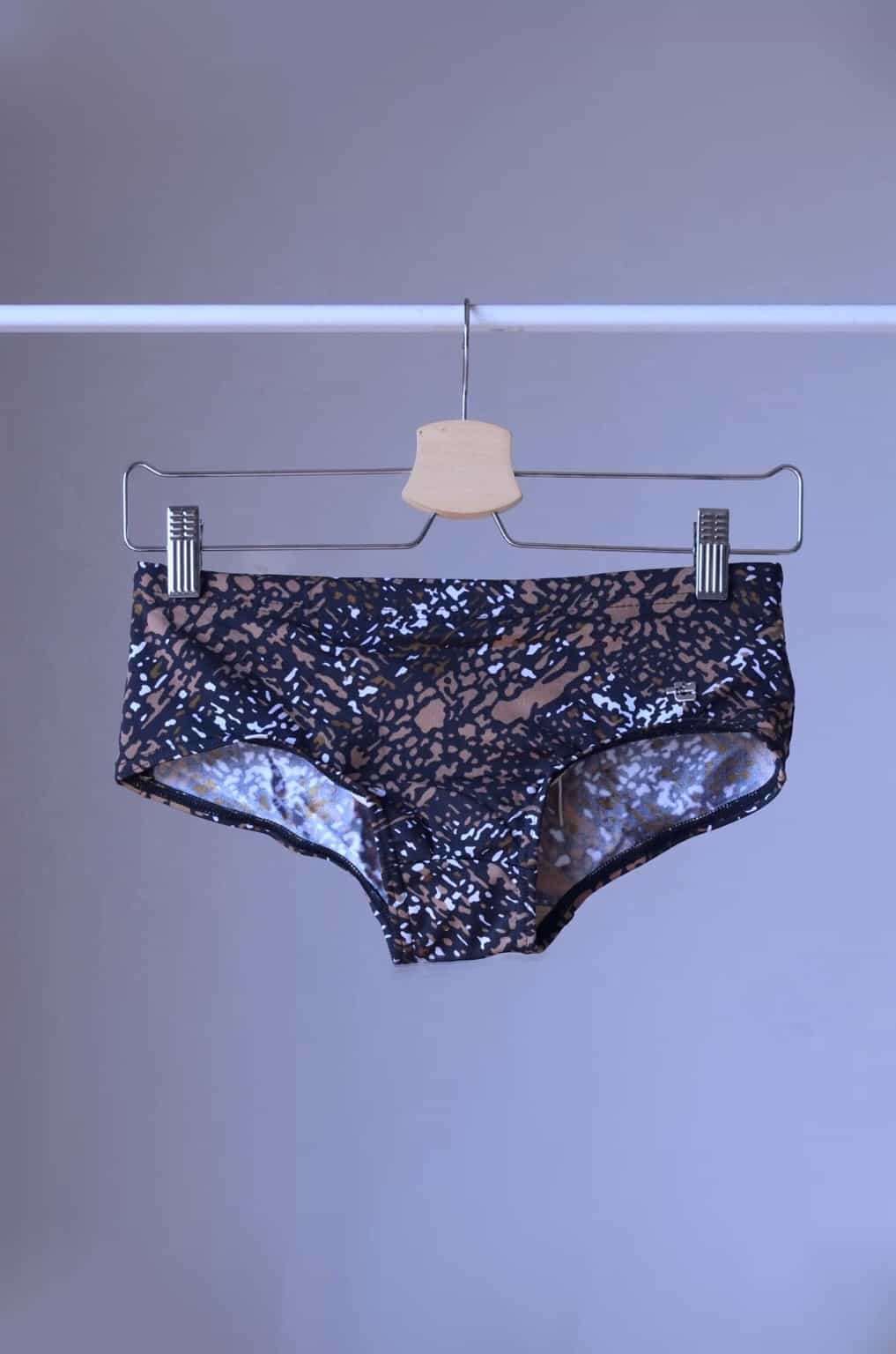Brown swim briefs with an abstract leopard print pattern, hanging on a wooden hanger. The pattern includes shades of brown, black, and white.