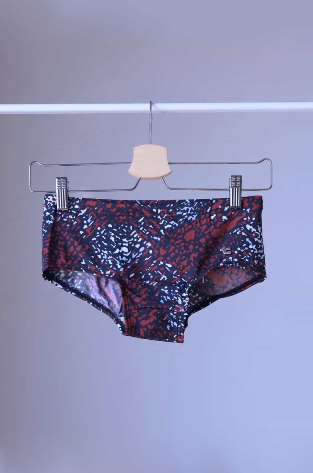 Maroon swim briefs with an abstract leopard print pattern, hanging on a wooden hanger. The pattern includes shades of brown, black, and white.