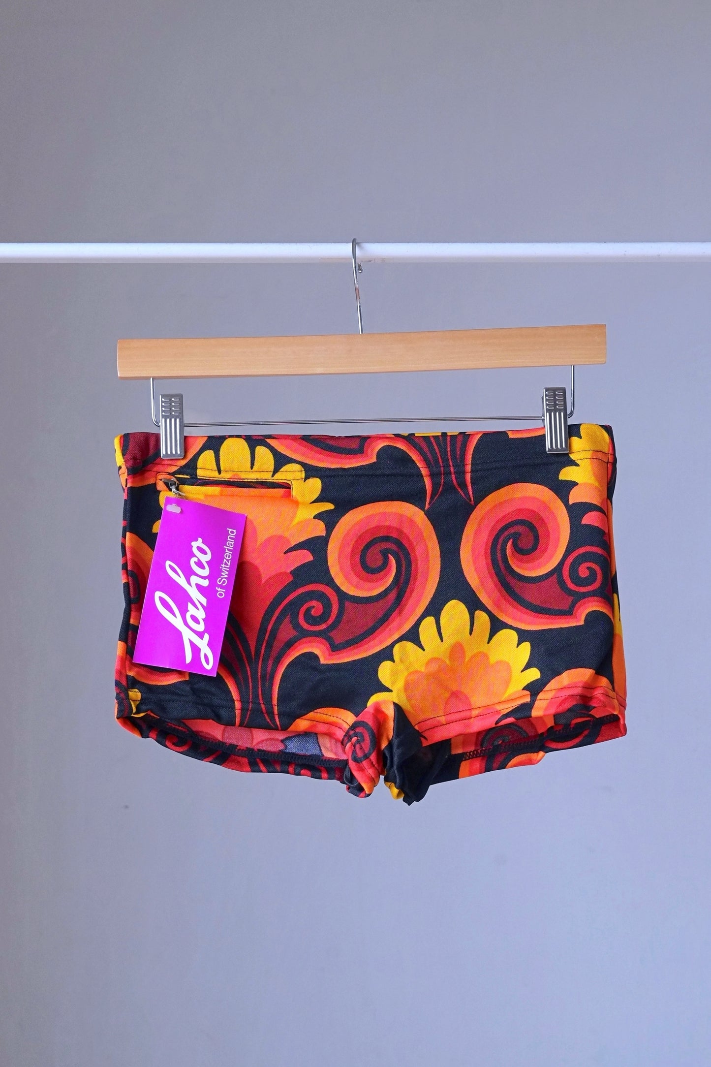 LAHCO vintage 60s square-cut swim trunks in red, yellow, and black psychedelic print with original tag, hanging on a wooden hanger.
