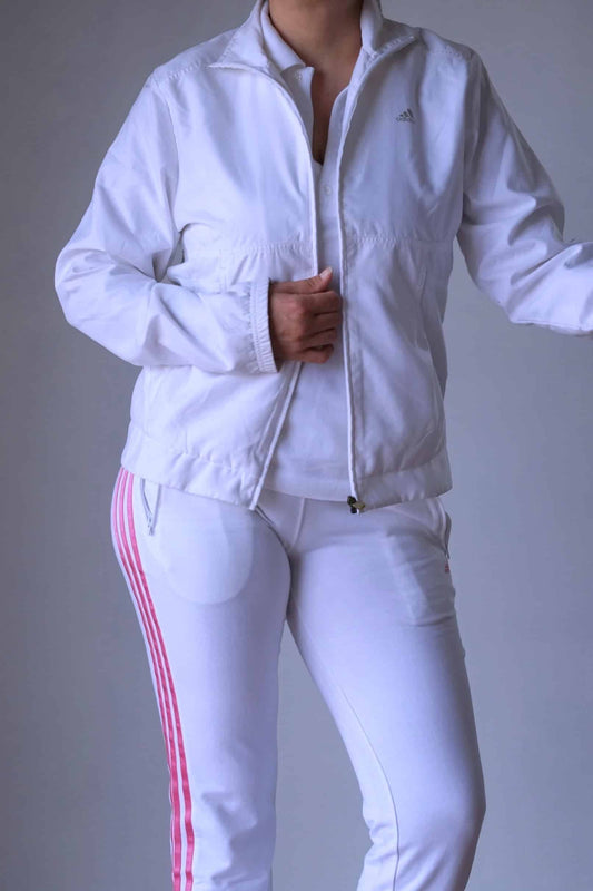 A person wearing a vintage 2000s Adidas women's white windbreaker jacket with a stand collar and full-zip front, paired with white pants featuring pink stripes down the sides. The jacket includes two side pockets and is styled with a classic, sporty look.