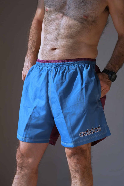 Man wearing vintage ADIDAS swim shorts with a blue front, purple back, and white stitched logo, standing with hands in pockets.