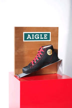 Angled view of vintage Aigle boot display. Wood back with logo, metal ramp, black canvas shoe with red laces on red base.