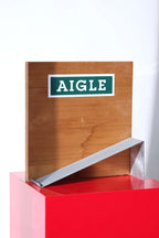 Empty vintage Aigle boot display stand. Wood backboard with green Aigle logo, metal boot ramp.