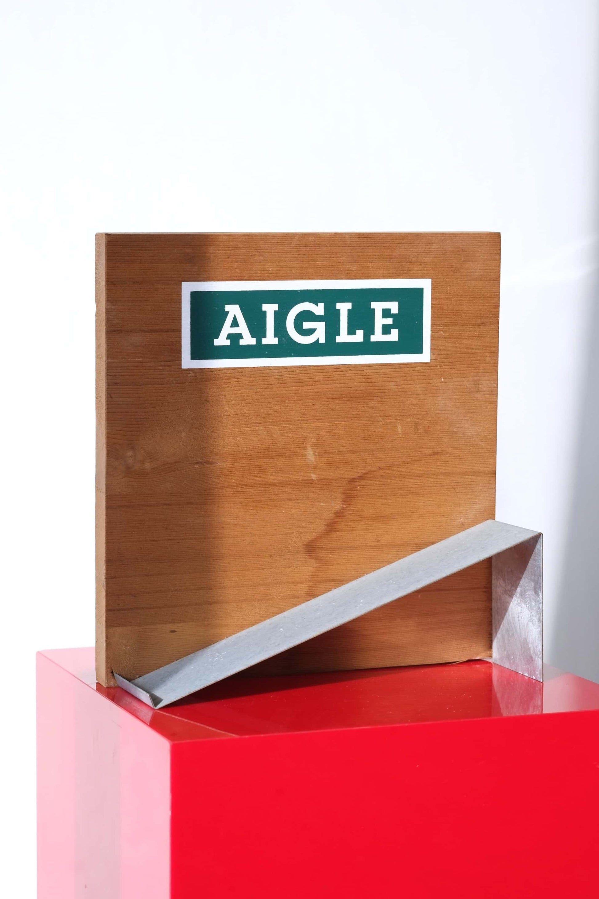 Empty vintage Aigle boot display stand. Wood backboard with green Aigle logo, metal boot ramp.