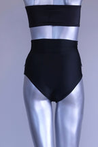 Back view of A pair of black, high-rise dance briefs with a folded-over waistband and a logo on the left hip, displayed on a mannequin.