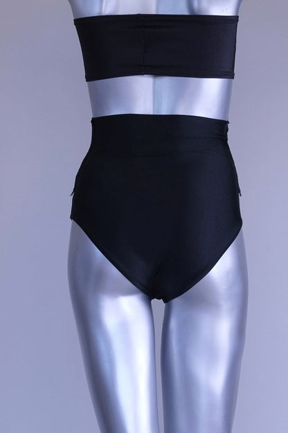 Back view of A pair of black, high-rise dance briefs with a folded-over waistband and a logo on the left hip, displayed on a mannequin.