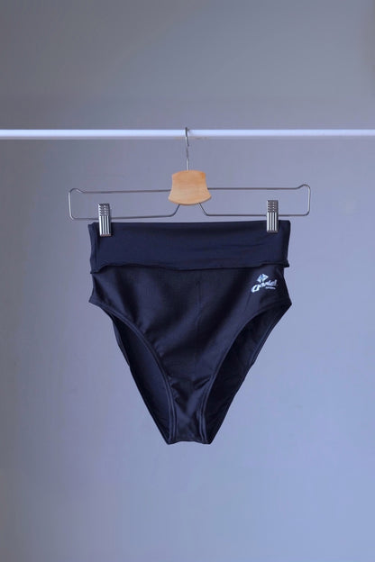 A pair of black, high-rise dance briefs with a folded-over waistband and a logo on the left hip, hanging on a metal hanger against a gray background.
