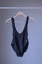 A black, vintage suspender leotard with a deep scoop neck and low back, hanging on a wooden hanger against a gray background.