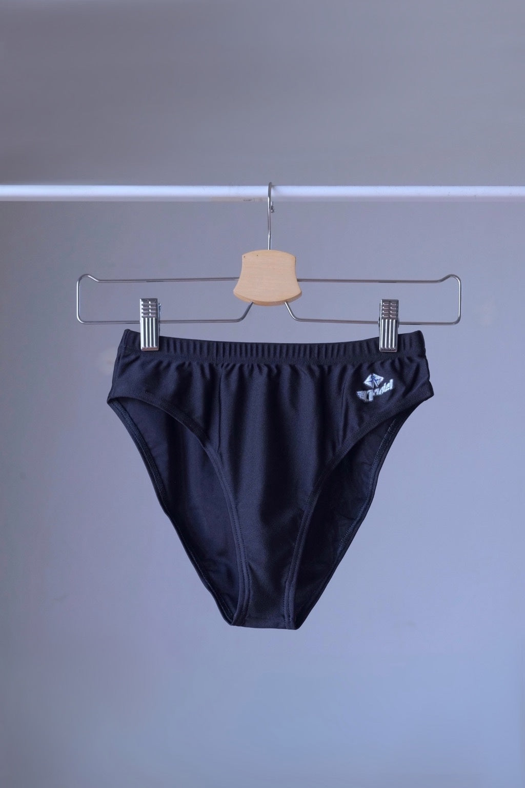 A pair of black, mid-rise dance briefs with a small logo on the left hip, hanging on a metal hanger against a gray background. The elastic waistband is visible.
