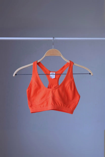 A vibrant orange vintage 90s ANNIEL sports bra with a racerback and elastic underbust band, hanging on a wooden hanger against a gray background.