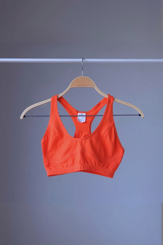 A vibrant orange vintage 90s ANNIEL sports bra with a racerback and elastic underbust band, hanging on a wooden hanger against a gray background.