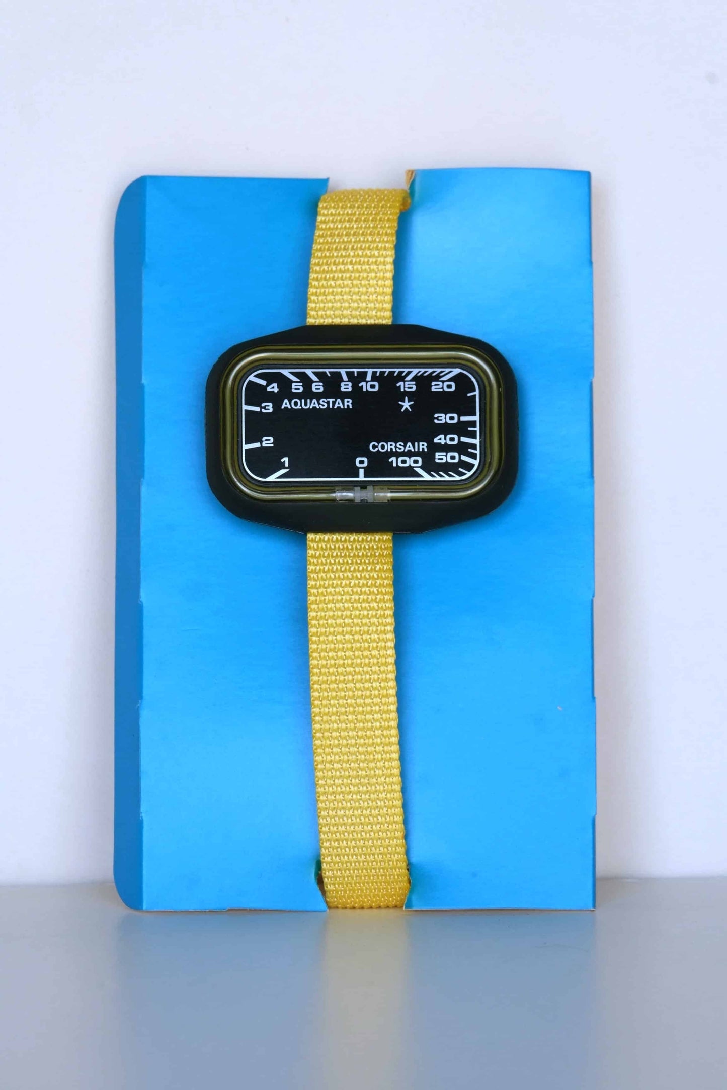 A vintage Aquastar Corsair analog depth gauge with a black rectangular dial, encased in a black frame. The depth gauge is attached to a vibrant yellow band and is displayed against a blue background. The dial features white markings and numbers, measuring depths up to 100 meters.