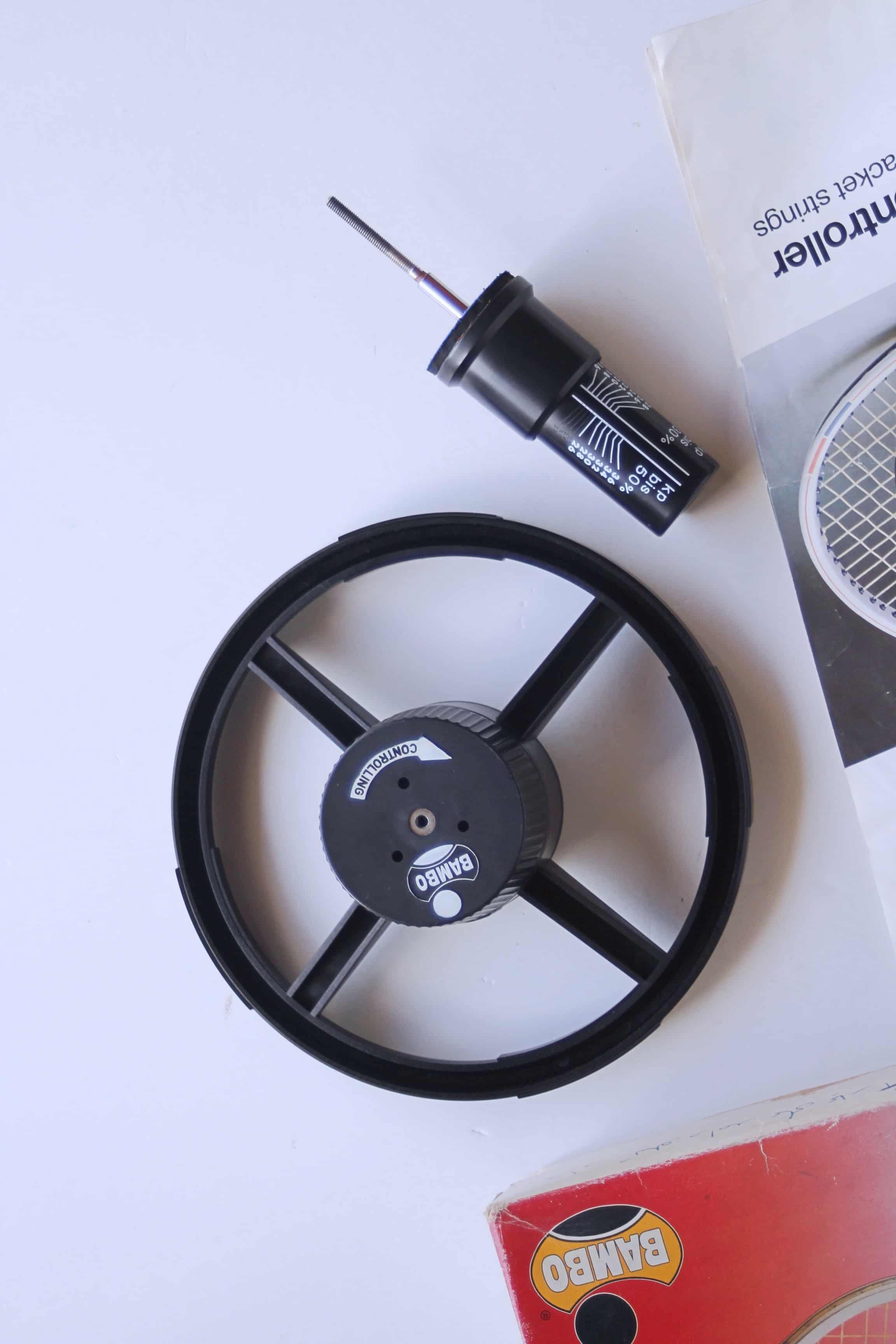Close-up view of the black BAMBO mechanical string tension tester wheel and its accompanying calibrated metal pin gauge tool.