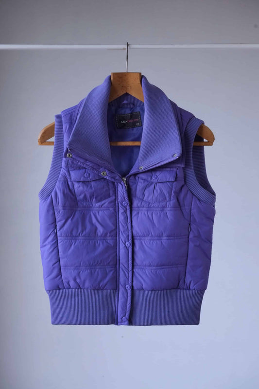 A short, sleeveless Y2K puffer vest in a vibrant shade of purple. It has a high collar and a zipper closure. The vest is hanging on a wooden hanger against a light gray background.