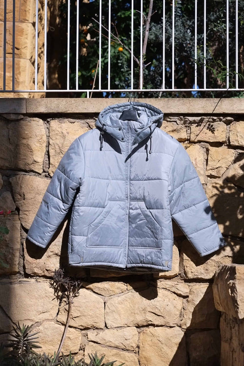 Brugi Y2K light grey hooded puffer jacket, full front view with horizontal quilting, zip closure and kangaroo pockets, photographed outdoors against stone wall.