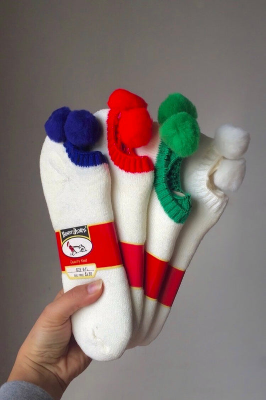Hand holding 4 pairs of pom pom socks, 1 blue/white pair, 1 red/white pair, 1 green/white pair and 1 white/white pair