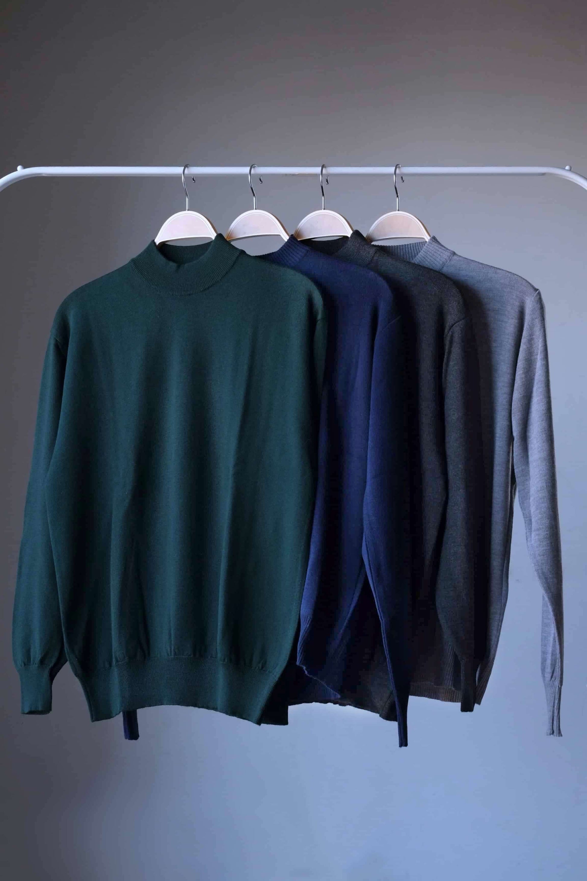 Vintage 90s Cagi pure wool high-neck sweaters in four colors (dark green, navy, heather charcoal, heather grey) hanging on a rail.