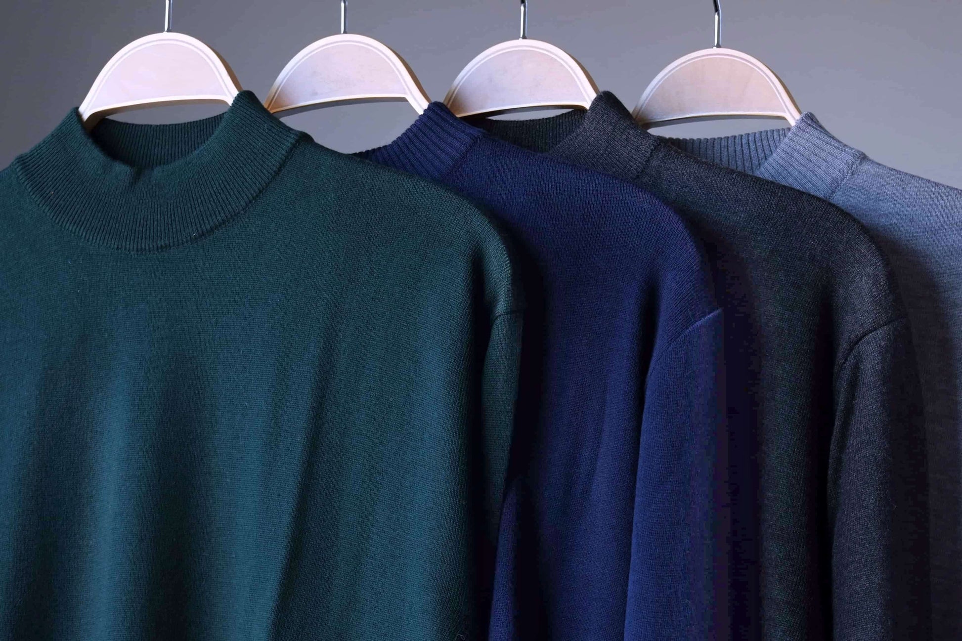 Tight shot showing the high-neck collars of the four Cagi Italian pure wool sweaters (green, navy, charcoal, grey). Note the mix of ribbed and unribbed neck finishes.