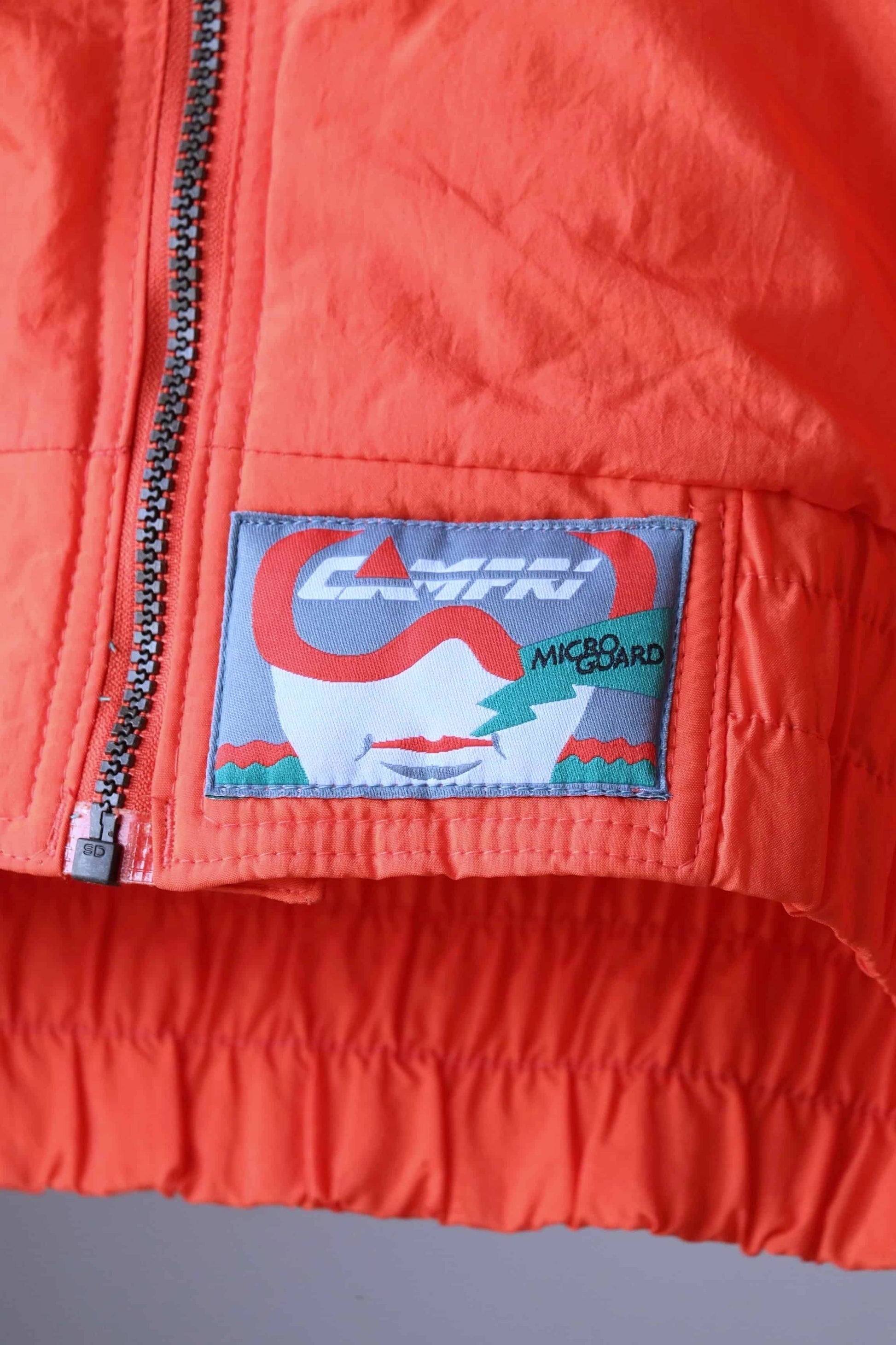 Close-up of a vintage orange jacket with a "CAMPRI" label and a smaller "MICROGUARD" patch. The label features a skier wearing goggles and a helmet.