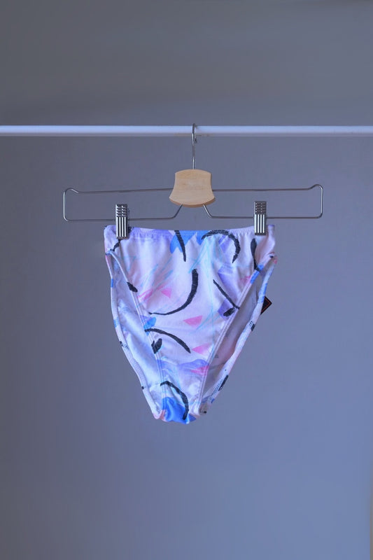 A pair of vintage 90s DANSKIN dance briefs with a white base and blue, purple, and black pastel print, displayed on a hanger.