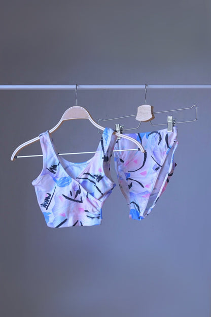 A DANSKIN vintage 90s matching activewear set, featuring a pastel abstract print crop top and high-cut briefs, displayed on hangers.