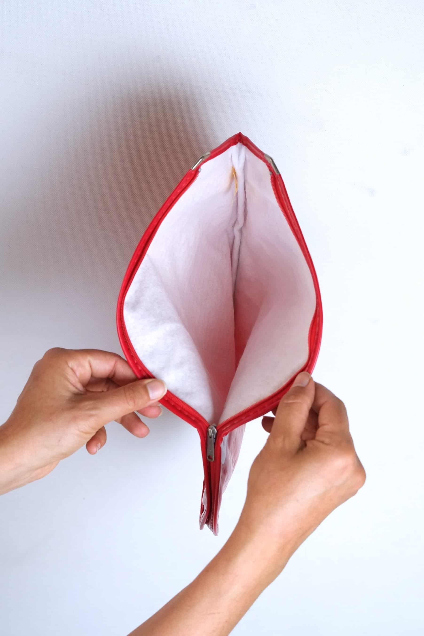 Two hands holding open a vintage red vinyl Dunlop tennis racket head cover, showing the soft white protective fleece lining and a silver metal zipper pull.