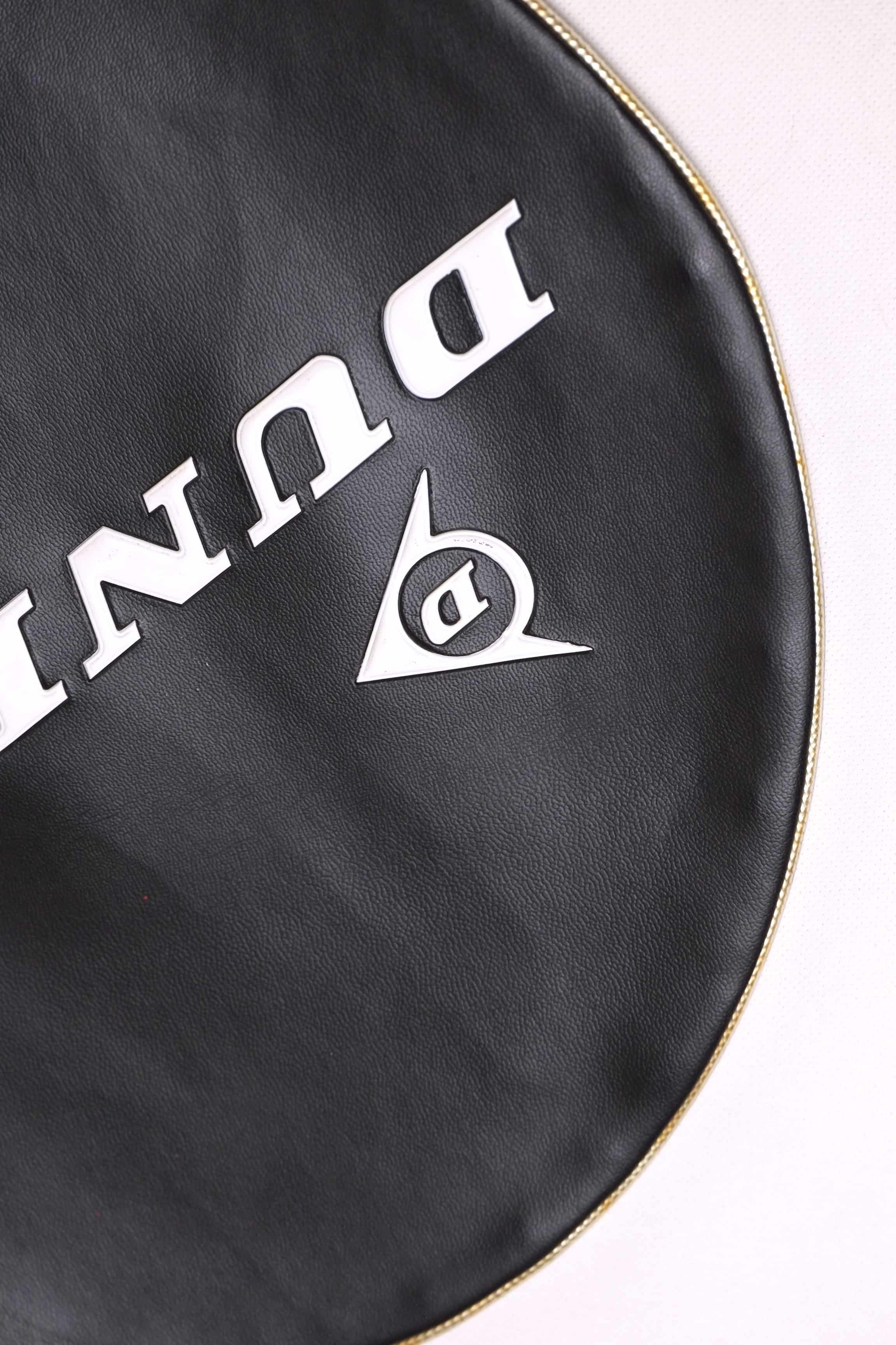 Detailed shot of the white embossed Dunlop logo and lettering on a black grained vinyl vintage tennis racket cover.