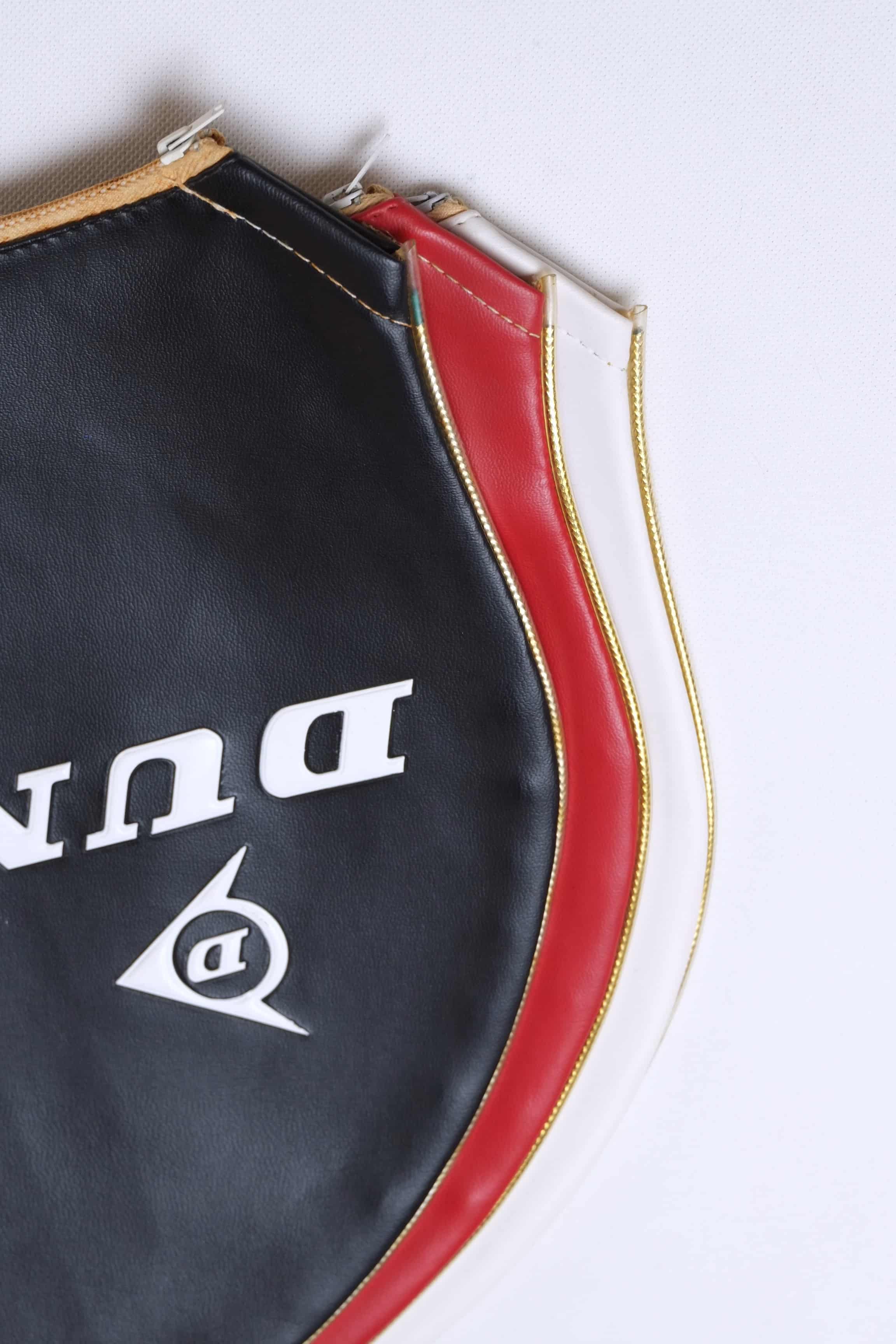 Close-up view of stacked vintage vinyl Dunlop tennis racket covers in black, red, and white, highlighting the stitched edges and gold-tone zippers.