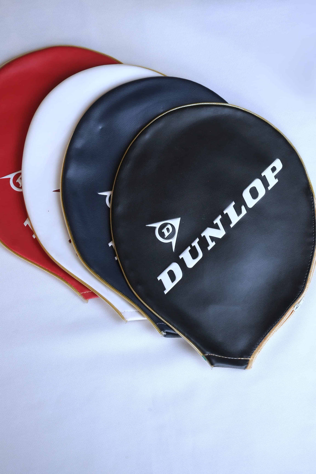 Three vintage vinyl Dunlop tennis racket head covers fanned out in red, white, navy and black with embossed white branding and gold-toned zipper trims, isolated on a white background.