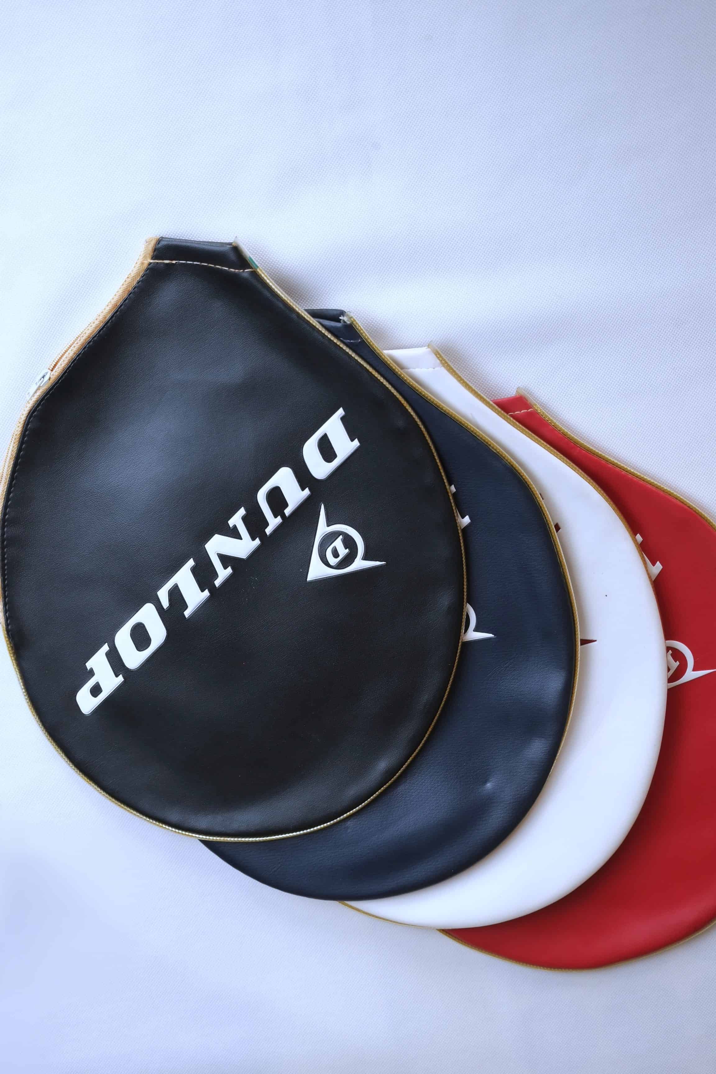 Three vintage vinyl Dunlop tennis racket head covers fanned out in red, white, navy and black with embossed white branding and gold-toned zipper trims, isolated on a white background.
