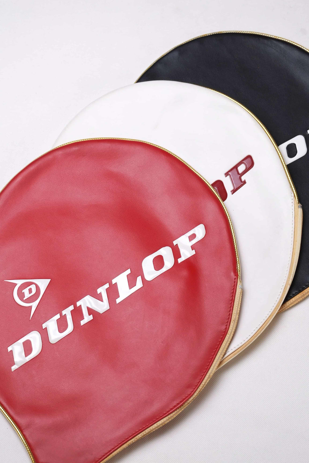 Overhead tight shot of three vintage Dunlop vinyl tennis racket head covers fanned out in red, white, and black with embossed white branding.