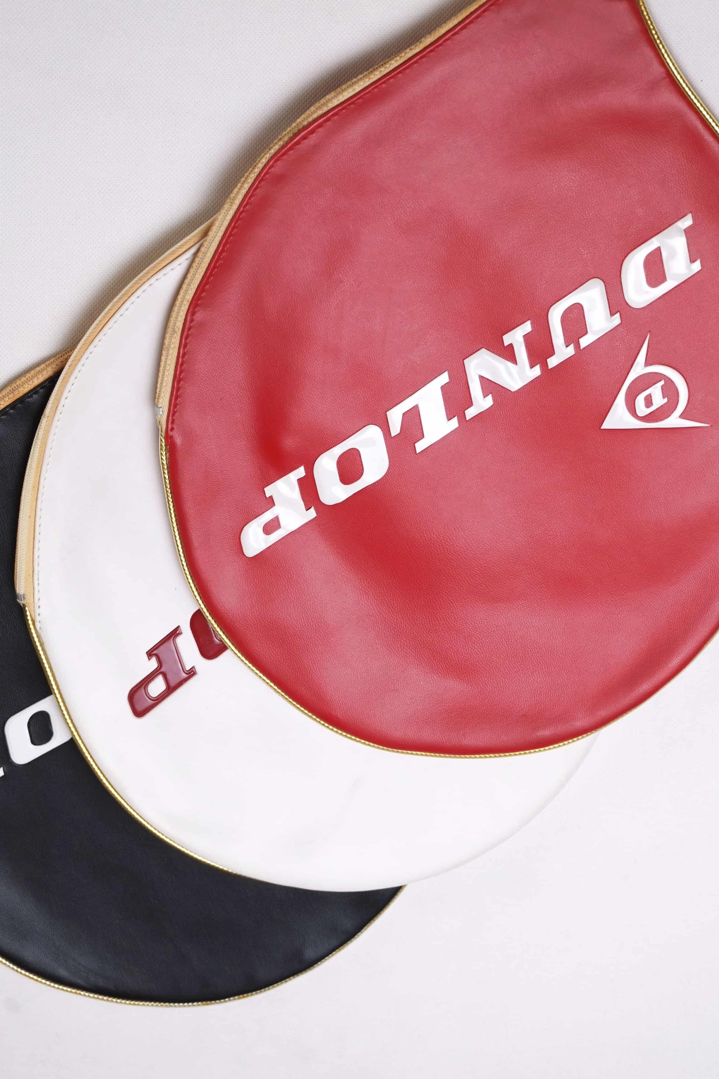 Overhead tight shot of three vintage Dunlop vinyl tennis racket head covers fanned out in red, white, and black with embossed white branding.