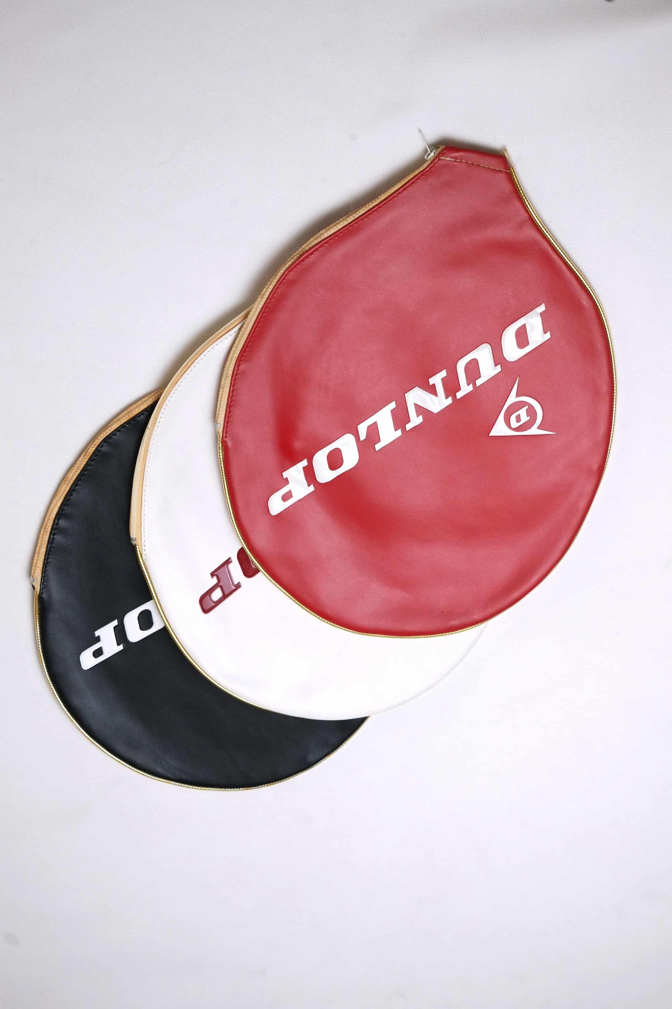 Three vintage vinyl Dunlop tennis racket head covers fanned out in red, white, and black with embossed white branding and gold-toned zipper trims, isolated on a white background.