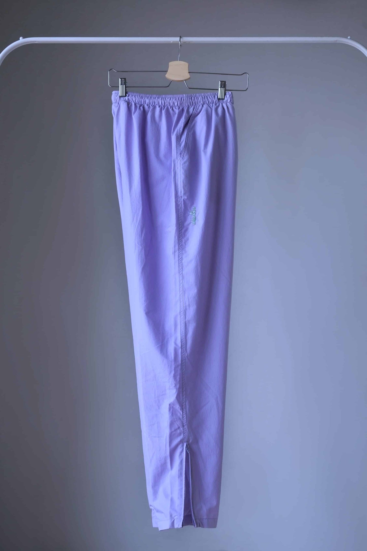 A pair of Vintage Ellesse track pants in a lilac shade,
featuring an elastic waistband and ankle zips, displayed on a hanger.