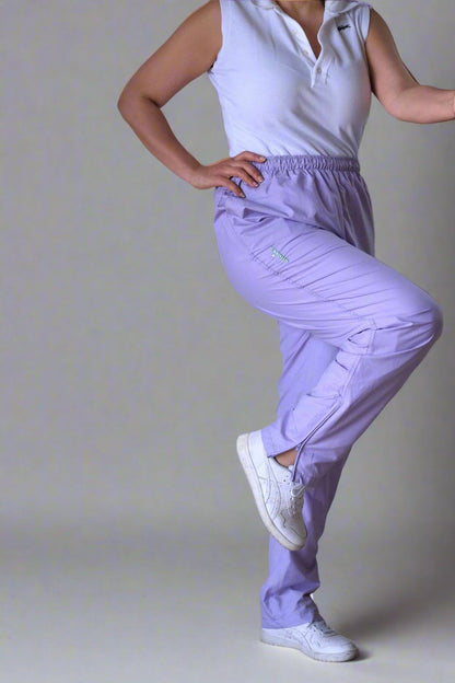 Person wearing vintage Ellesse lilac track pants with an elastic waistband and relaxed fit, paired with a sleeveless white polo shirt and white sneakers, standing indoors against a neutral background. The person is flexing their right leg at the knee.